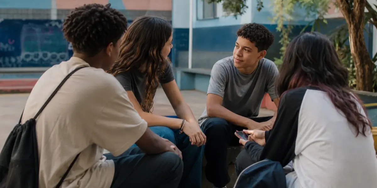 Grupo diverso de adolescentes conversa em um espaço comunitário ao ar livre, com um celular aparecendo discretamente.
