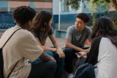 Grupo diverso de adolescentes conversa em um espaço comunitário ao ar livre, com um celular aparecendo discretamente.