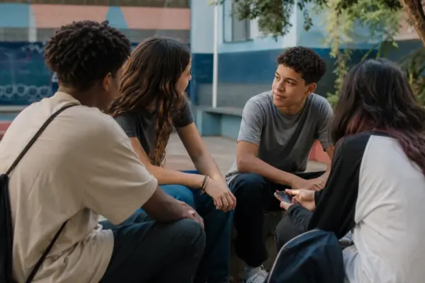 Grupo diverso de adolescentes conversa em um espaço comunitário ao ar livre, com um celular aparecendo discretamente.