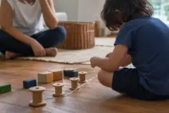 Criança vista de costas organiza carretéis, blocos e fios no chão enquanto uma pessoa adulta acompanha ao fundo.