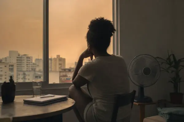 Pessoa anônima observa uma cidade sob céu enevoado de calor através de uma janela, com ventilador e caderno ao lado.